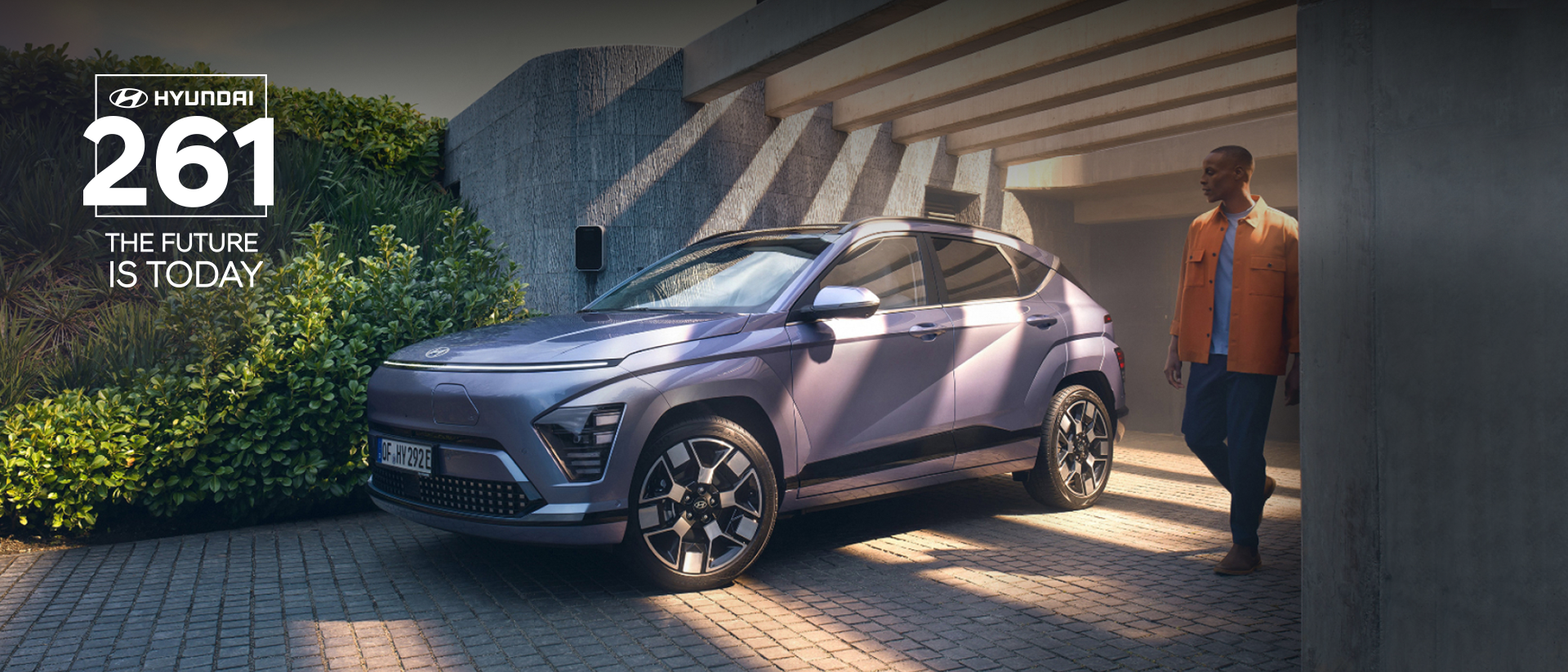 A white Hyundai Kona Electric car is parked with a man standing on the left side and a woman walking past on the right side. The background features a modern building with large windows.